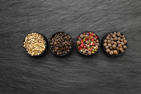 Black, green, pink, white and fragrant pepper grains in round jars placed on black stone surface backgroundの写真素材