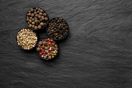 Black, green, pink, white and fragrant pepper grains in round jars placed on black stone surface backgroundの写真素材