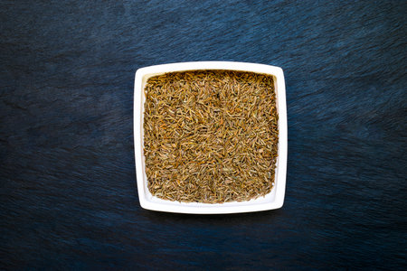 Dry caraway seeds in white plate placed on deep blue and black stone background surface with free...
