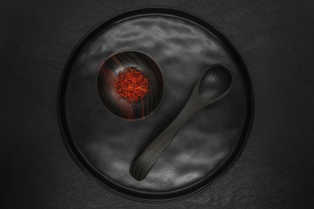 Composition of Saffron in black wooden bowl with black wooden spoon on black round plate and black stone backgroundの写真素材