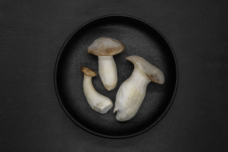 Abstract composition of three Eringi mushrooms in black cast iron bowl on grey stone backgroundの写真素材