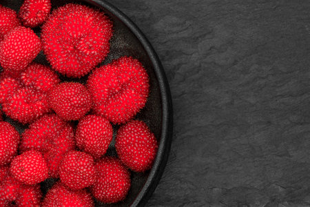 Red balloon berries (Rubus Illecebrosus) in round and black iron cast bowl on black stone background...