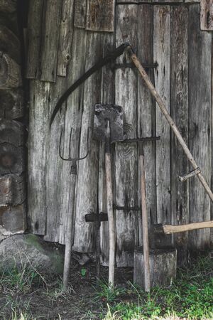 Very old, rusty and dirty garden working rural tools (hoe, scythe, spade, rake, ax, pitchfork) near the barn wooden wall in countryside during summer timeの写真素材