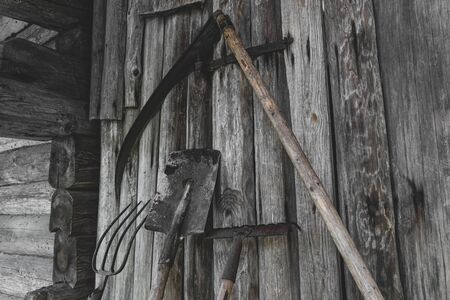 Very old, rusty and dirty garden working rural tools (scythe, spade, rake, pitchfork) near the barn wooden wall in countryside during summer timeの写真素材