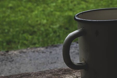 Part of very old with a lot of scratches white metallic mug in front of the green grass background nd placed on wooden plank with copy spaceの写真素材