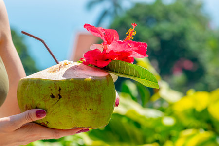 Woman holding fresh green coconut drink cocktail with red and white flowers in front of the blue and...