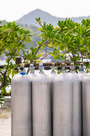 A lot of aluminum scuba diving oxygen tanks standing in front of the green beach bushes and sea gulf on the sand in Thailand with copy spaceの写真素材