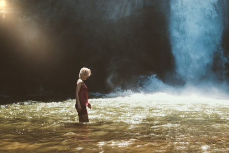 Young blonde woman with pink blouse and shorts walking in brown water river and illuminated by the...