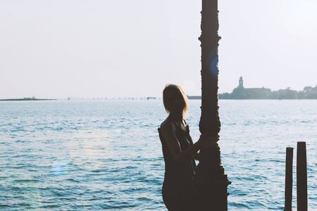 Travel and leisure concept. Dark woman silhouette in blue sea canal background in Venice, Italy and illuminated by sun beams and lens flares. Vintage tone filter color style with high exposureの写真素材