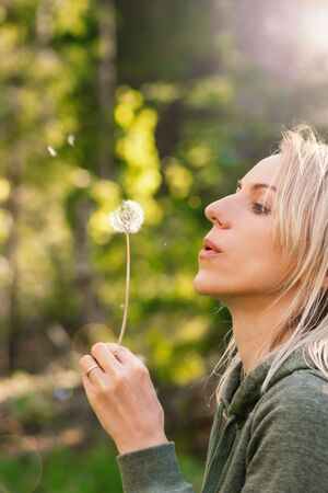 Blonde woman blowing dandelion blossom fluffs in the background of blurred dense green forest during summer evening time and illuminated by sun beams and lens flareの写真素材