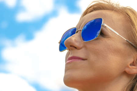 Blonde womans close-up profile face with blue sunglasses and pink lipstick in blue sky with white clouds background with copy space