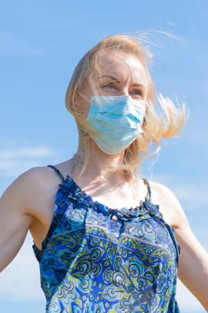 Healthcare and medicine concept. Portrait of blonde woman with windswept hair and medicine mask on her face. Woman wear blue dress in blue sky background with copy spaceの写真素材