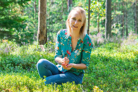 Blonde woman with colorful blouse collecting cranberries in dense green forest and illuminated by summer sun beamsの写真素材