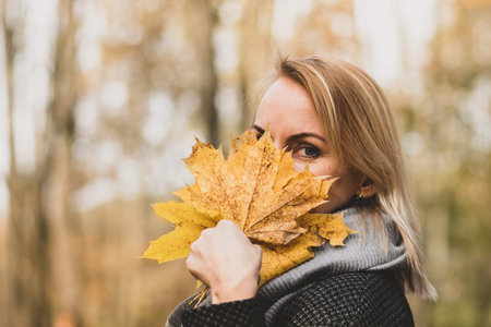 Autumn concept. Blonde woman cover face with yellow maple tree leaves bouquet and watching with one...