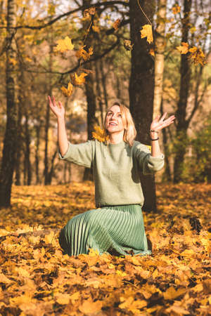 Autumn concept. Blonde woman with green blouse and skirt catching yellow falling maple tree leaves in forest during sunny autumn dayの写真素材
