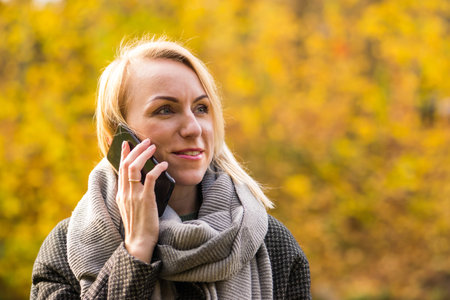 Business and autumn concept. Blonde woman with grey coat speaking with smartphone in front of the yellow autumn nature with copy spaceの写真素材