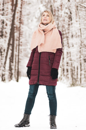 Portrait of blonde woman with purple coat and pink scarf standing in the forest during winter time and watching natureの写真素材