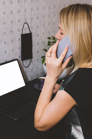 Healthcare concept. Blonde woman working with computer with white blank screen for copy space and speaking with cell phone from home during quarantine. Woman sitting at the desk with coffee cup and hanging black medicine mask on the wall with copy space. Hand is in camera focusの写真素材