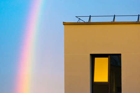 Rainbow in the blue evening sky and part of flat apartment with window illuminated by yellow sunset lightの写真素材
