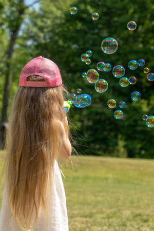 Little girl with pink hat and long hairs blowing soap bubbles in air in front of the forest background. Girl standing back to cameraの写真素材