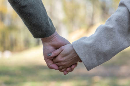 Love, relationship, nature concept. Close-up shot of man and woman holds each other hands in blurred nature background during cold and sunny dayの写真素材