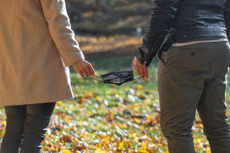 Close-up view from back of man and woman holds black medicine face mask as a symbol of breaking or connecting pandemic relationships in blurred nature background during cold and sunny autumn dayの写真素材