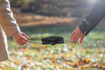 Close-up view from back of man and woman holds black medicine face mask as a symbol of breaking or connecting pandemic relationships in blurred nature background during cold and sunny autumn dayの写真素材