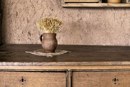 Abstract rural composition of clay vase with a bouquet of dried cereals standing on old, grunge and aged wooden desk with drawers in front of clay wall with wooden shelf and copy space. HDR imageの写真素材