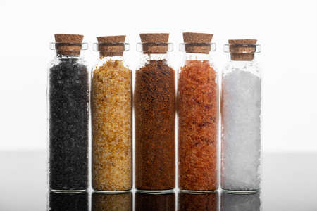 Food concept. Various spices in cylinder shape glass bottles with cork in white background standing on dark reflecting surface. Various colorful salt types. Studio shotの写真素材