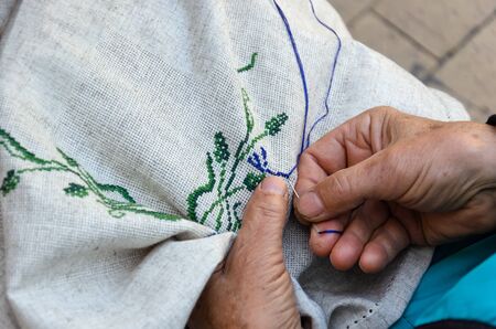 Hands of an elderly woman embroidering a cross-stitch floral pattern on linen fabric. Embroidery, handwork, needlecraft concept. Close-upの写真素材