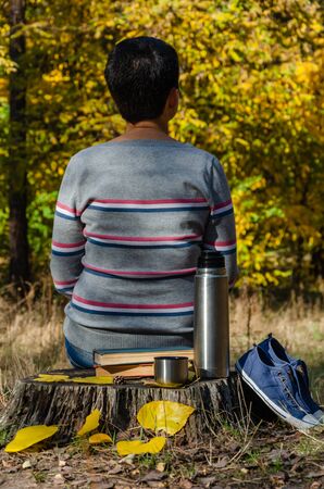 A middle-aged or young woman, dressed in casual clothes, sitting on the stump in the autumn park. Books, flask bottle and a cup of coffee or tea, laceless denim sneakers. Calm concept. Back viewの写真素材