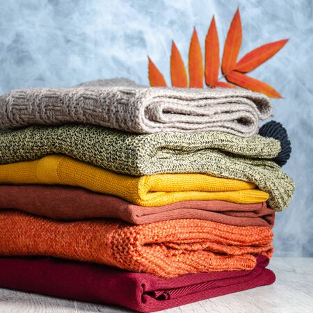 Stack of folded warm knitted women's sweaters in warm colors and bright autumn leaves on gray background. Copy space. Closeupの写真素材