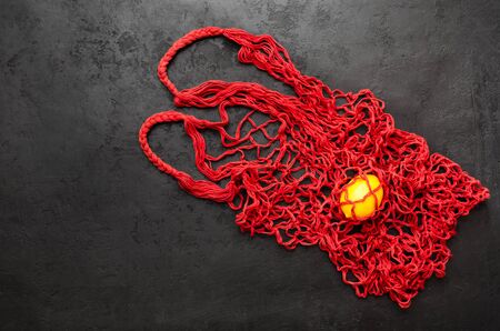 Trendy mesh net bag - alternative to plastic bags. Single yellow lemon in red reusable string bag on black background. Zero waste shopping concept. Eco friendly lifestyle. Top view. Copy spaceの写真素材