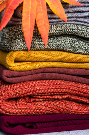 Stack of folded warm knitted women's sweaters in warm colors and bright autumn leaves. Close-up