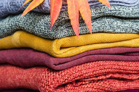 Stack of folded warm knitted women's sweaters in warm colors and bright autumn leaves. Close-up