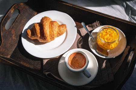 Weekend breakfast in bed. Wooden tray with coffee, fresh croissant and orange jam on linen in bright...