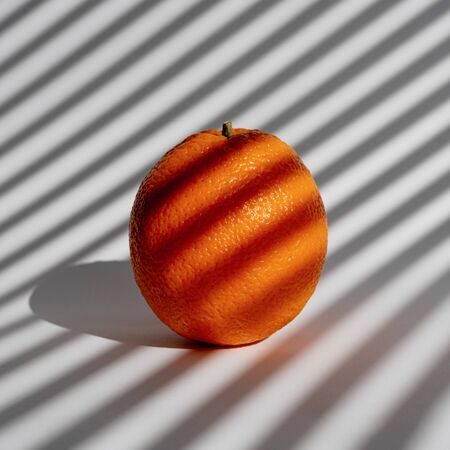 Ripe juicy orange on a white background in morning light through the louvres. Streaks of sunlight from the window blinds. Creative minimal designの写真素材