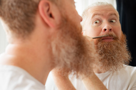 Attractive middle-aged man cutting his moustache and beard by himself in front of mirror at home. Beard care during quarantine. Coronavirus outbreak, lockdown, staying home on self-isolation. Closeupの写真素材