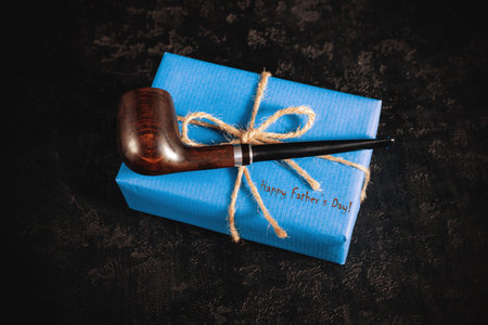 Blue gift box and smoking tobacco pipe on black background. Happy father's day concept. Greeting...