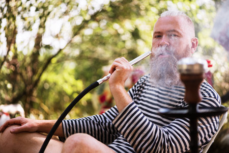 Bearded man smoking hookah outdoors. Hipster dressed in striped sailor shirt exhaling smoke....