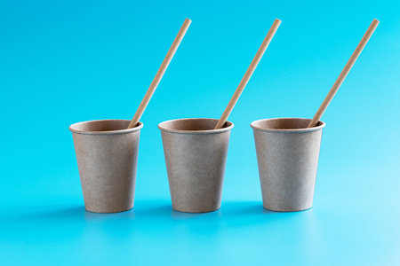 Three disposable brown craft paper cups with paper straws in a row on blue background. Eco-friendly...