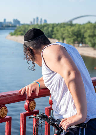 Unrecognizable young jewish man wearing twisted payot in black velvet kippah standing on the bridge and looking down into river. Vertical orientationの写真素材