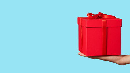 Hand holding red gift box with ribbon bow isolated on light blue background. Holiday concept. Copy spaceの写真素材