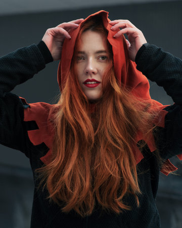 Portrait of beautiful young woman with red long hair in hood looking at camera. Girl in the image of Red womanの写真素材