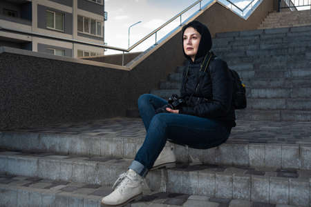 Confident woman photographer holding camera in her hands sitting on steps outdoors. Beautiful mid aged woman in hood looking into the distance.の写真素材