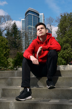 Handsome confident teenage boy sitting on the stairs in the city park on spring sunny day. Relaxed teenager dressed red hoodie and black jeans dreaming outdoors enjoying lifeの写真素材