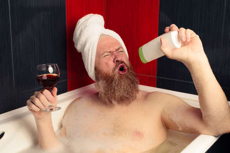 Cheerful bearded man taking bubble bath holding glass of red wine and singing into shampoo bottle with facial expression. Relaxation at home after hard day. Happy guy enjoying lifeの写真素材