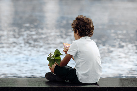 Unrecognizable curly boy holding ice cream cone and green branch in his hands, sitting in city park contemplating water in fountain and dreaming. Rear viewの写真素材