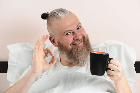 Happy bearded man enjoying morning coffee in bed showing OK gesture. Smiling hipster guy starting...