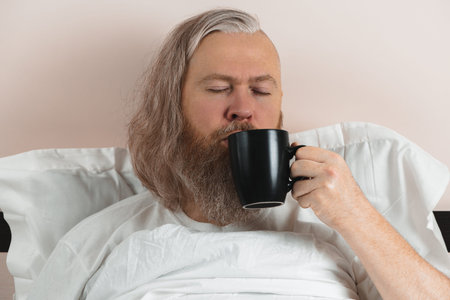 Bearded man enjoying his morning coffee in bed to cheer up before starting new day. Closeup